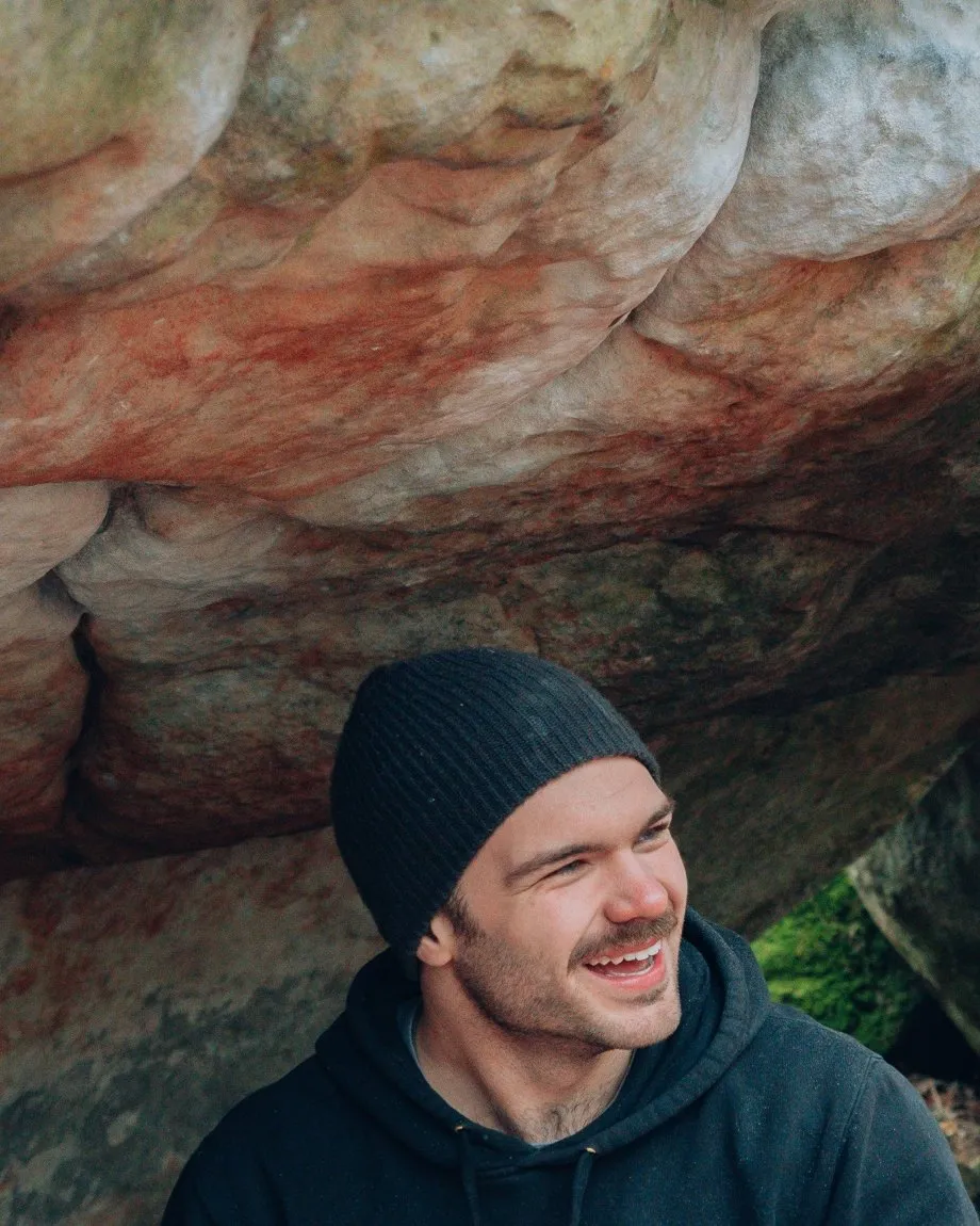 Frank Blokdijk smiling, sitting under an orange-streaked sandstone roof in Fontainebleau, in a black beanie and hoodie.